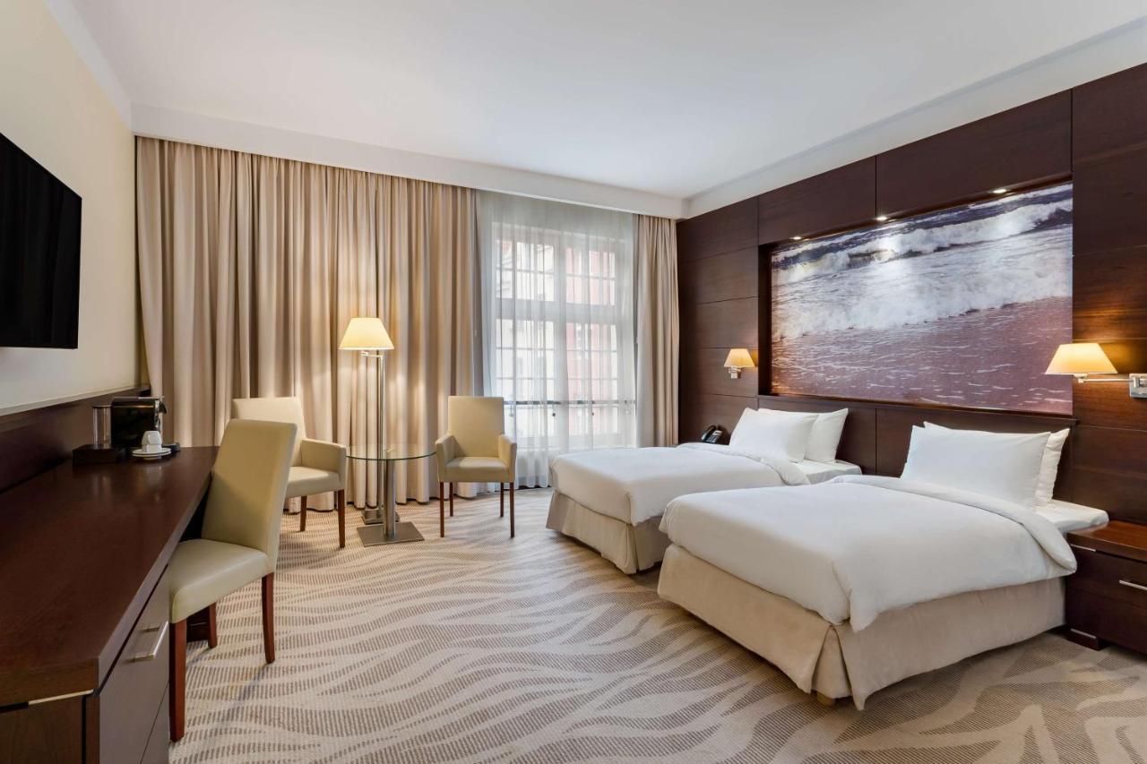 Radisson Blu Hotel, Gdansk Bagian luar foto The photo shows a neatly arranged hotel room featuring two beds with white bedding. There is a large window covered with light curtains, allowing natural light to enter the space. In one corner, there are two chairs and a small table, creating a cozy