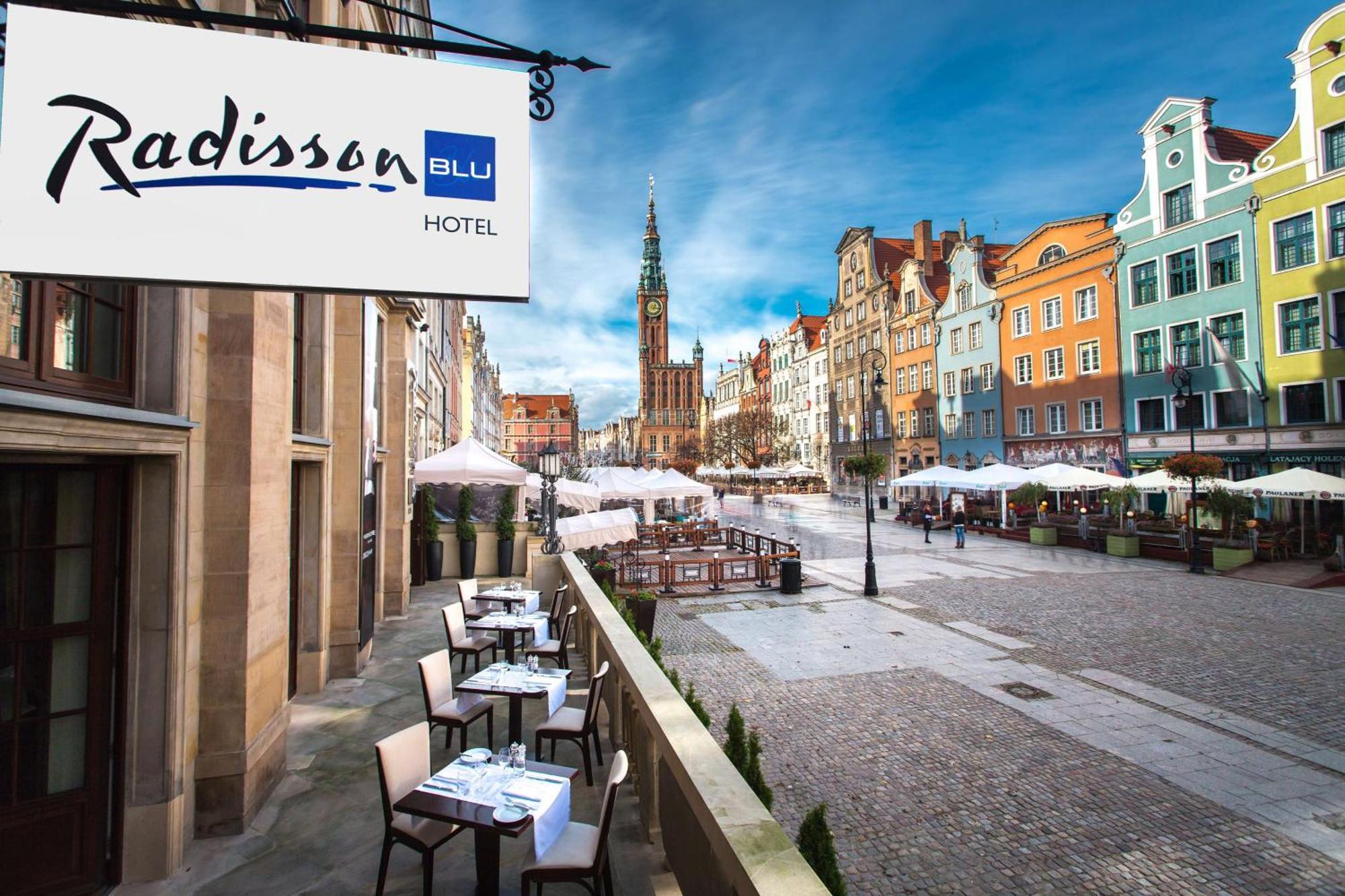 Radisson Blu Hotel, Gdansk Bagian luar foto The photo shows a view of the Radisson Blu Hotel, which is situated in a vibrant, picturesque area. The image features an outdoor seating arrangement with tables and chairs, likely part of a restaurant or café. In the background, colorful historic bu
